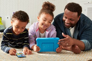 An image of a dad and two kids laying on a carpet smiling at their devices, including a tablet and a phone. 